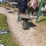 Ženska potiska samokolnico po poti na dvorišču, druga oseba pa stoji ob njej.