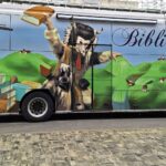 Bibliobus z napisom »Bibliobus« in poslikavo knjig, gričev ter kurenta stoji ob robu ceste.