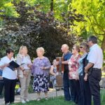Pevci ob spremljavi harmonike nastopajo na vrtu doma starejših pred zbranimi stanovalci.