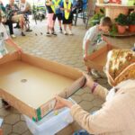 Otroci in starejša oseba sodelujejo pri igri z veliko kartonsko podlago in žogicami.