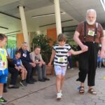 Deklica in starejši moški hodita po označeni poti, medtem ko ju opazujejo otroci in odrasli.