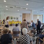 Stanovalci sedijo v večnamenskem prostoru in spremljajo skupino otrok v rumenih majicah.