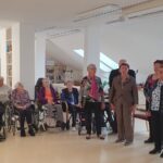 Pevska skupina nastopa v skupnem prostoru doma, medtem ko stanovalci, sedeči na stolih in invalidskih vozičkih, spremljajo nastop.