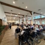 V večji dvorani doma za starejše številni stanovalci sedijo na stolih in invalidskih vozičkih ter spremljajo nastop pred njimi.