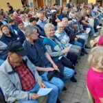 Stanovalci doma spremljajo gledališko predstavo na prostem med številnim občinstvom.