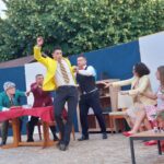 Igralci na odru uprizarjajo komični prizor med gledališko predstavo na prostem.