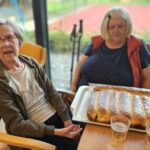 Dve stanovalki sedita za mizo ob pečeni potici in kozarcih soka.