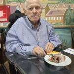 Oseba v invalidskem vozičku sedi za mizo s kosom torte na krožniku.