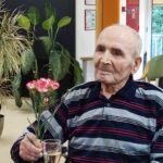 Oseba sedi za mizo s kozarcem penine in v roki drži šopek nageljnov.
