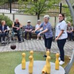 Skupina stanovalcev spremlja met krogle proti kegljem na zunanjem igrišču.