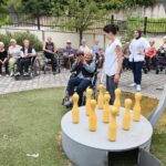 Stanovalka v invalidskem vozičku drži kroglo ob igrišču za rusko kegljanje, ob njej stojita spremljevalki, v ozadju pa sedi skupina stanovalcev.