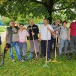 Skupina stanovalcev in zaposlenih pozira pod drevesom z vrtnim orodjem po skupni delovni akciji urejanja okolice.