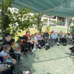 Skupina otrok in odraslih sedi pod pergolo, poraslo z vinsko trto.