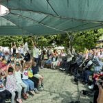 Otroci in odrasli pod pergolo sodelujejo pri skupni vadbi z dvignjenimi rokami.