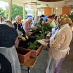 Skupina udeležencev sadi okrasne rastline v cvetlična korita na dolgi mizi pod nadstreškom.