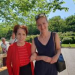 Stanovalka doma starejših in spremljevalka stojita skupaj na sončnem dvorišču med druženjem ob izletu.