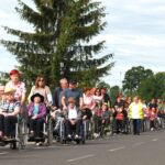 Skupina stanovalcev na invalidskih vozičkih in spremljevalcev se sprehaja po cesti skozi Malo vas.