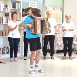 Mladostnik igra harmoniko, okoli stojijo ženske v belih majicah.