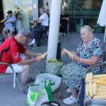 Stanovalca sedita in lupita koruzne storže poleg pladnja s koruzo pred stavbo.