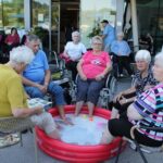 Skupina stanovalcev sedi z nogami v rdečem napihljivem bazenčku z milnico in barvnimi žogicami pred stavbo.