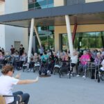 Stanovalci in osnovnošolci sedijo zunaj pred stavbo, ženska v beli majici vodi vajo z rokami.