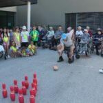 Stanovalec modri kapi meče žogo proti kegljicam, ženska drži rdeče krogle, stanovalci in otroci gledajo.