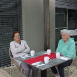 Dve stanovalki sedita za mizo na zunanji terasi z belimi skodelicami.