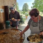 Stanovalec in stanovalka stojita ob velikem loncu z jedjo, stanovalka drži skledo s kruhom.