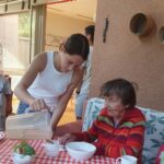 Oseba iz vrča naliva pijačo v skodelico pred sedečo žensko za mizo.