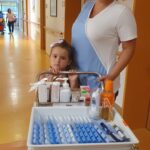 Ženska v delovni uniformi in deklica stojita za vozičkom z medicinskimi pripomočki.