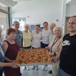 Skupina stanovalcev in osebja drži pladenj s fokačo, paradižniki in olivami.