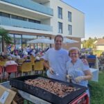 Moški in ženska stojita ob žaru, na katerem se pečejo čevapčiči, v ozadju sedi skupina ljudi.