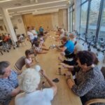 Udeleženci sedijo ob dolgi mizi in izdelujejo predmete iz volne in kartonskih tulcev.