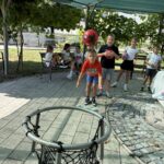 Skupina otrok se na prostem igra z žogami, eden od njih meče žogo proti košu.