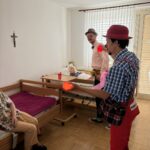 Dve osebi z rdečima nosovoma in klobukoma stojita ob posteljah, ena drži svetleče žogice.