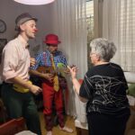 Dve osebi z rdečima nosovoma stojita ob ženski, ena igra ukulele.