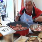 Moški v predpasniku s prijemalkama pripravlja narezane klobase ob posodi in kruhu.