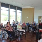 Skupina ljudi sedi v polkrogu in posluša govorko ob majhni mizi.