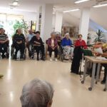 Moški s kitaro sedi pred skupino sedečih oseb v prostoru.