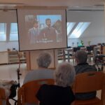 Skupina ljudi sedi pred projekcijskim platnom, na katerem se predvaja film s podnapisi.