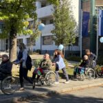 Skupina ljudi z modrimi baloni spremlja osebe na invalidskih vozičkih po pločniku.