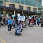 Skupina ljudi, nekateri na invalidskih vozičkih, se z modrimi baloni odpravlja izpred stavbe; v ospredju je napis »Sprehod za spomin, 21. 9., svetovni dan Alzheimerjeve bolezni«.