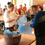Dve osebi pri mizi razvrščata modre kose blaga iz pletene košare, v ozadju sedi skupina ljudi.