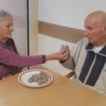 Dve osebi sedita ob mizi, ena drugi ponuja sladko kroglico s krožnika.