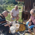 Tri osebe na prostem luščijo koruzo ob vedrih in kupu koruznih storžev.