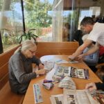 Stanovalke in moški sedijo za mizo ob oknih ter iz časopisnega papirja izdelujejo papirnate vrečke.