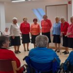 Skupina pevk nastopa pred sedečim občinstvom, ob strani sta notno stojalo in harmonika.