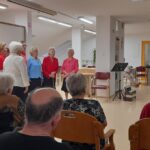 Skupina pevk stoji pred sedečim občinstvom in poje.