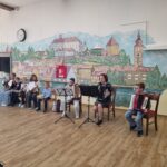 Skupina otrok in odrasla glasbenica sedi pred stensko poslikavo mesta ter nastopa z violinami in harmonikami.