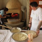 Ženska z dolgim pripomočkom ureja žerjavico v krušni peči, ob njej pa druga ženska v belem predpasniku pripravlja testo za gibanico.